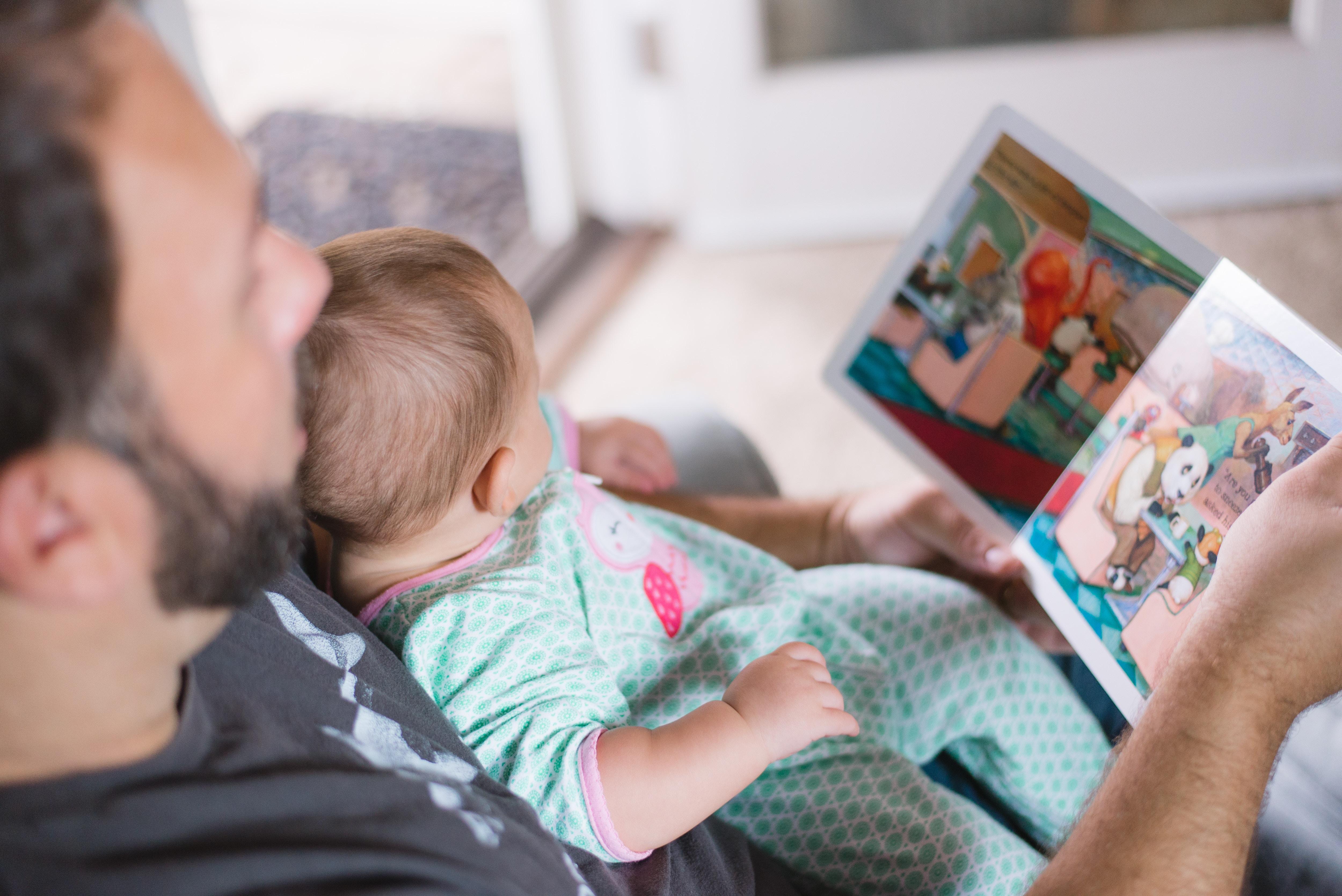 a male adult holds a baby on a lap while reading the baby a story