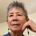 Elderly person with short gray hair and blue earrings thoughtfully resting their chin on their hand.