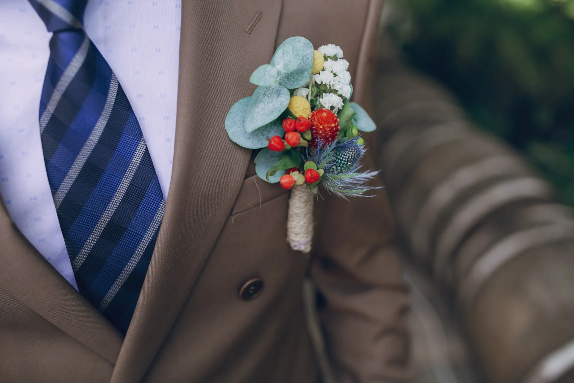 Man in brown suit with a colorful boutonniere pinned to his lapel, wearing a blue plaid tie.