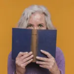 Woman in a purple sweater holds a book, partially covering her face against a vibrant orange background.