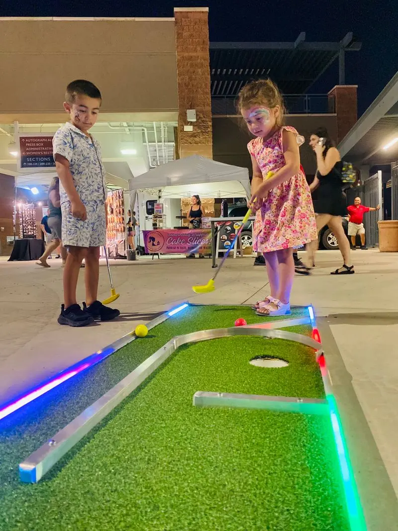 Two children play mini-golf with neon lights at an evening event. Stalls and people are visible in the background.